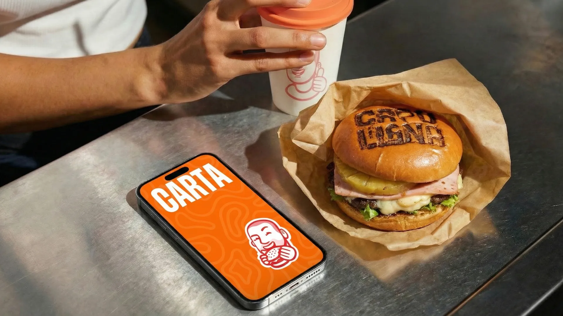Celular mostrando la carta naranja junto a una hamburguesa Capullana envuelta en papel y una mano sosteniendo un vaso sobre una mesa metálica