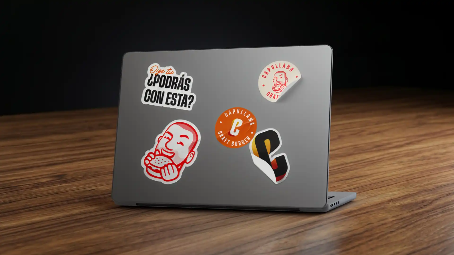 Laptop gris sobre una mesa de madera con varios stickers de la marca Capullana Craft Burger pegados en la tapa