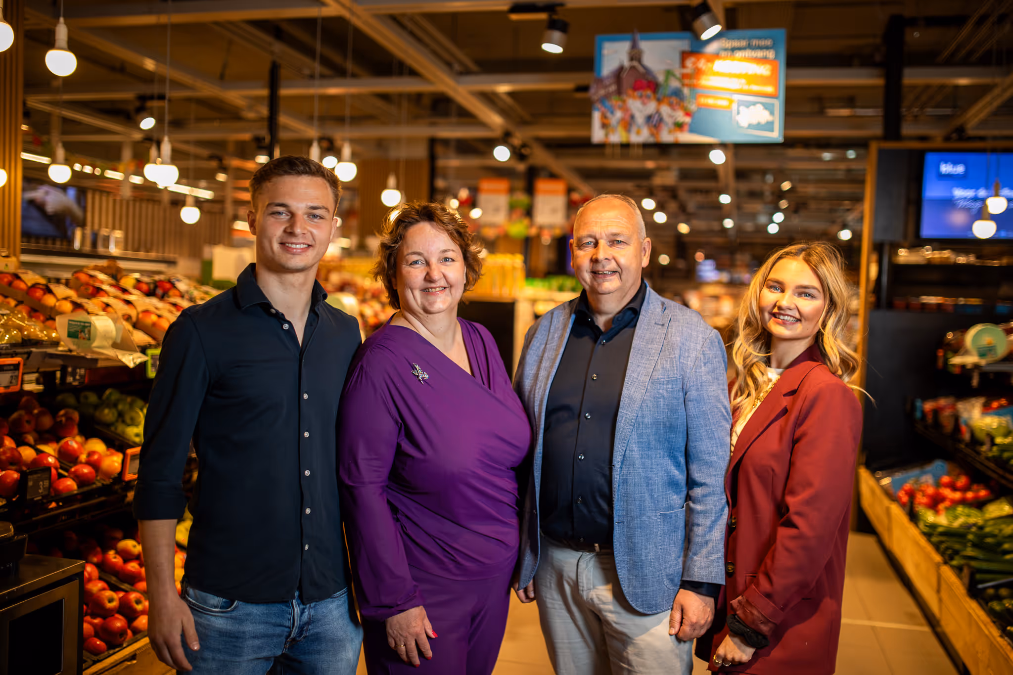 Vier lachende mensen poseren samen in een supermarktafdeling met groenten en fruit.