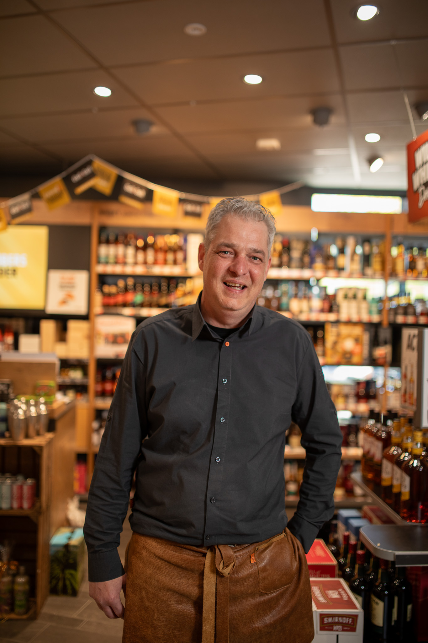 Gelukkige winkelmedewerker met grijs haar en leren schort staat in een winkel met gedistilleerde dranken.