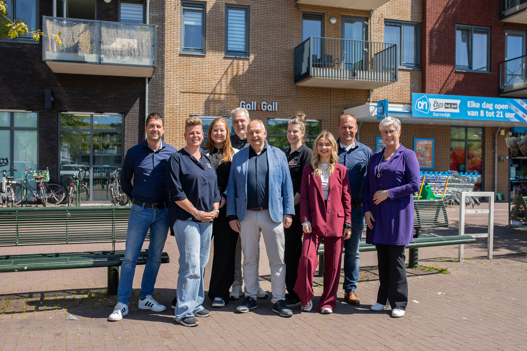 Groep van negen mensen poseren buiten voor een winkelpand met borden van Gall & Gall en Albert Heijn.