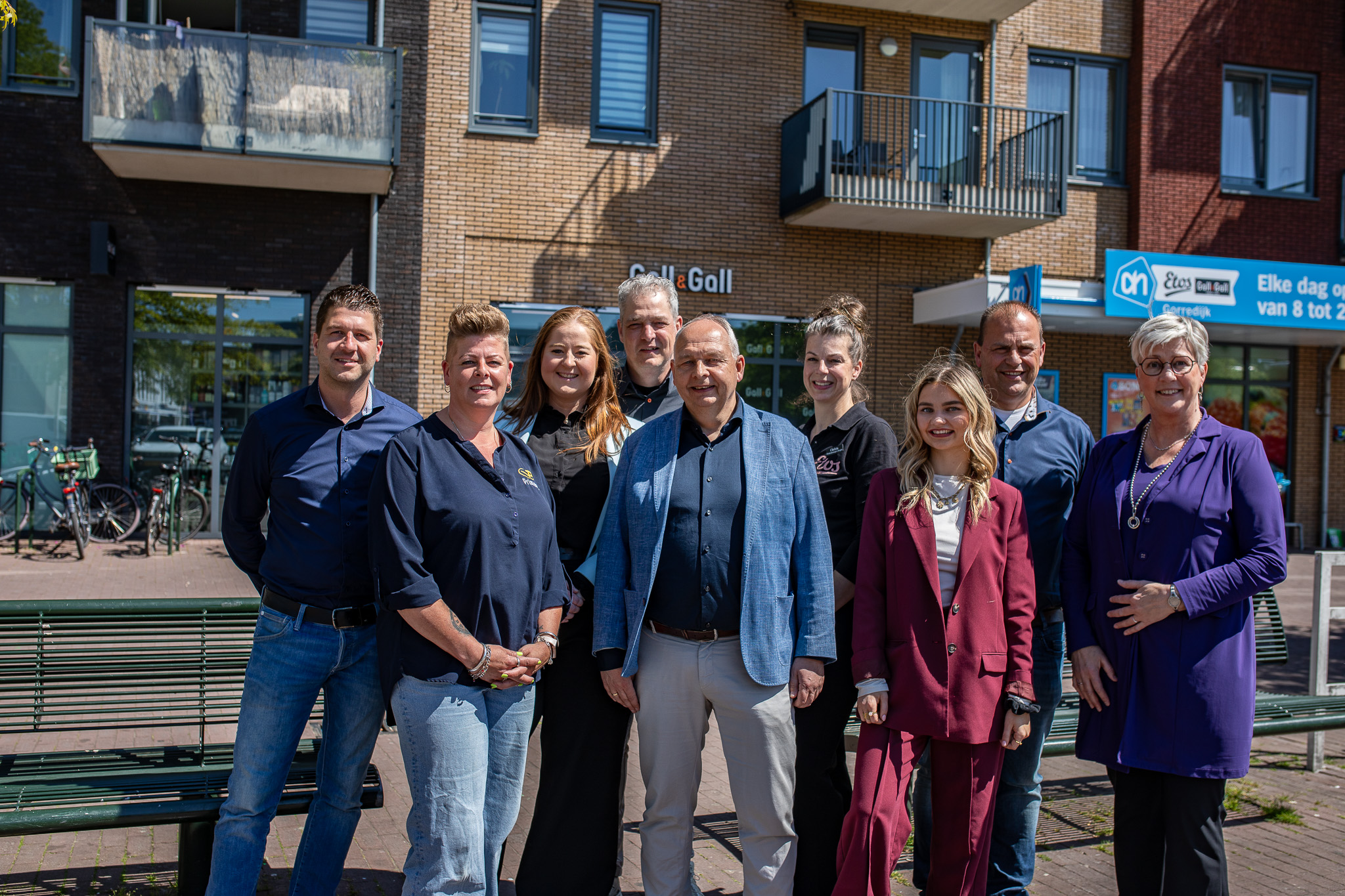 Groep van negen mensen staan samen buiten voor een winkelpand met fietsen en bankjes.