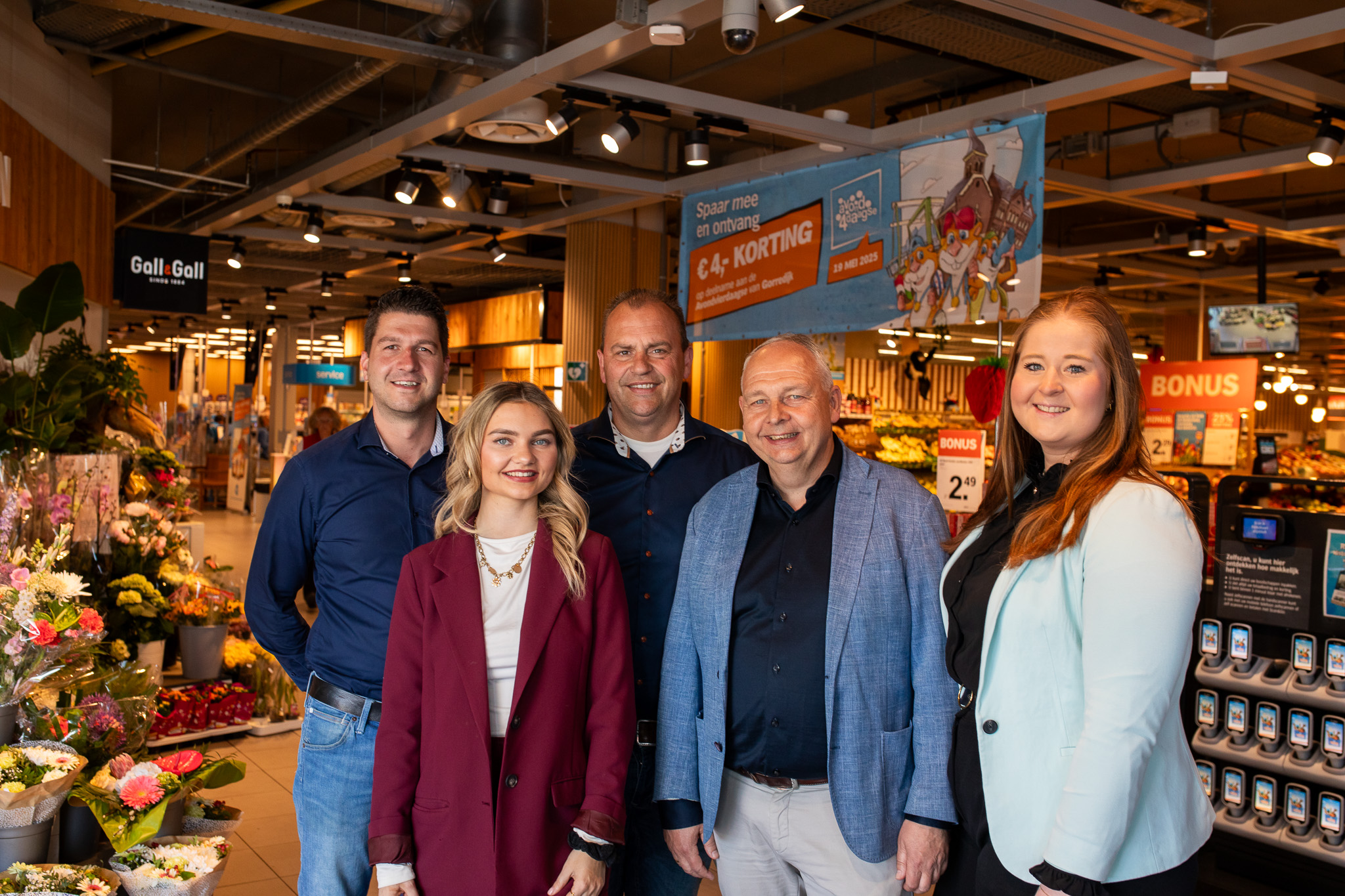 Vijf glimlachende medewerkers poseren in een supermarkt bij bloemen en promotieborden.
