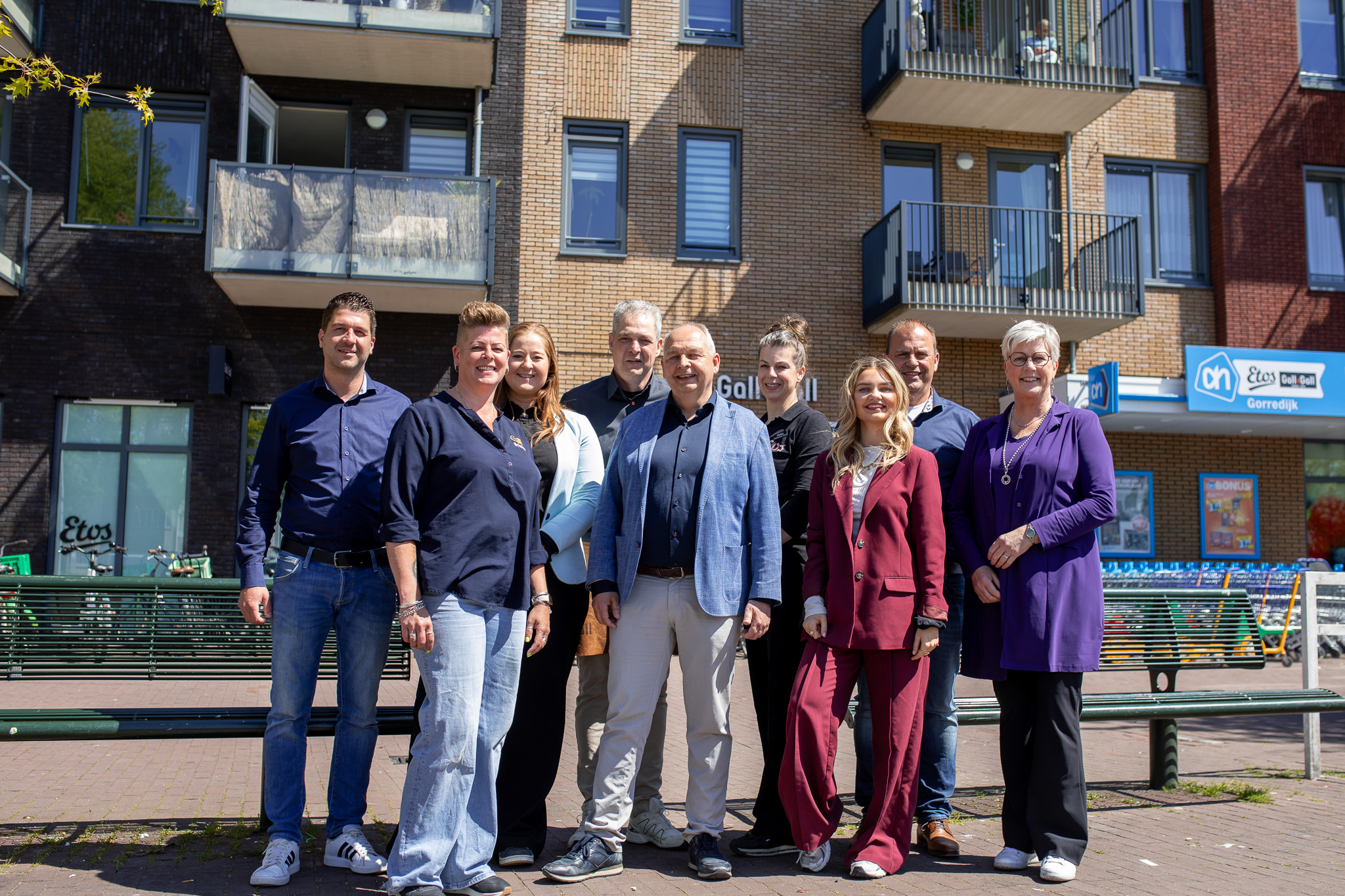 Groep van negen mensen die buiten voor een supermarkt en appartementencomplex poseren op een zonnige dag.