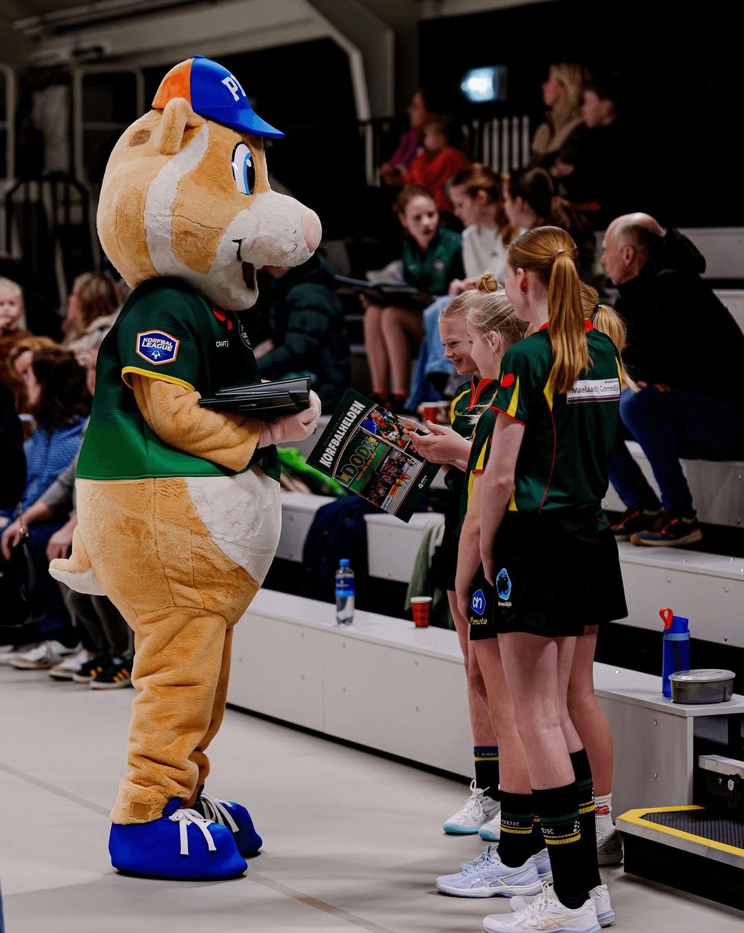 Mascotte met blauw petje en groene trui praat met drie jonge korfballers in groene sportkleding in een sporthal.