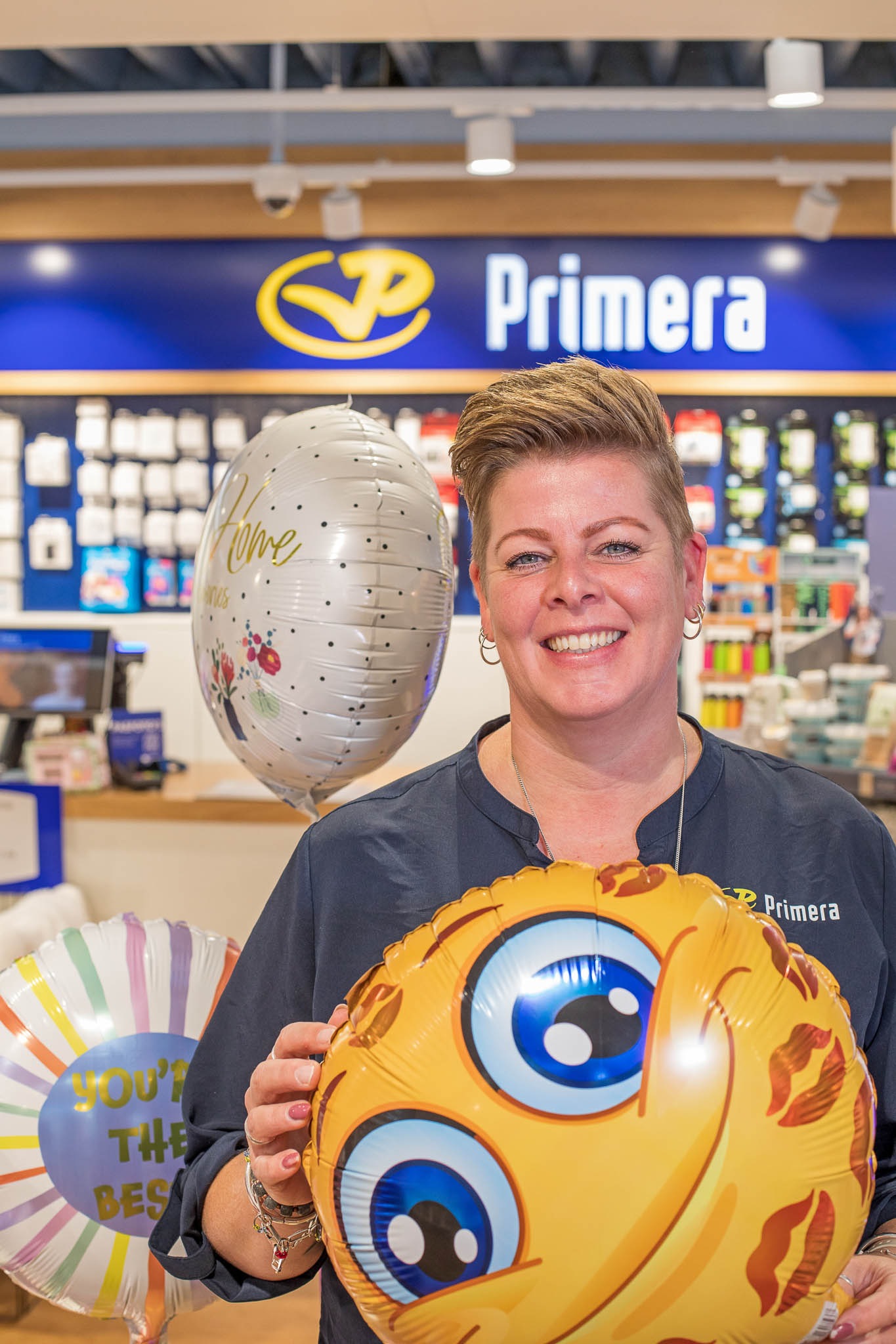 Vrouw in Primera-winkel lacht en houdt een ballon met een grote glimlachende emoji met blauwe ogen vast, met daarachter twee andere ballonnen.