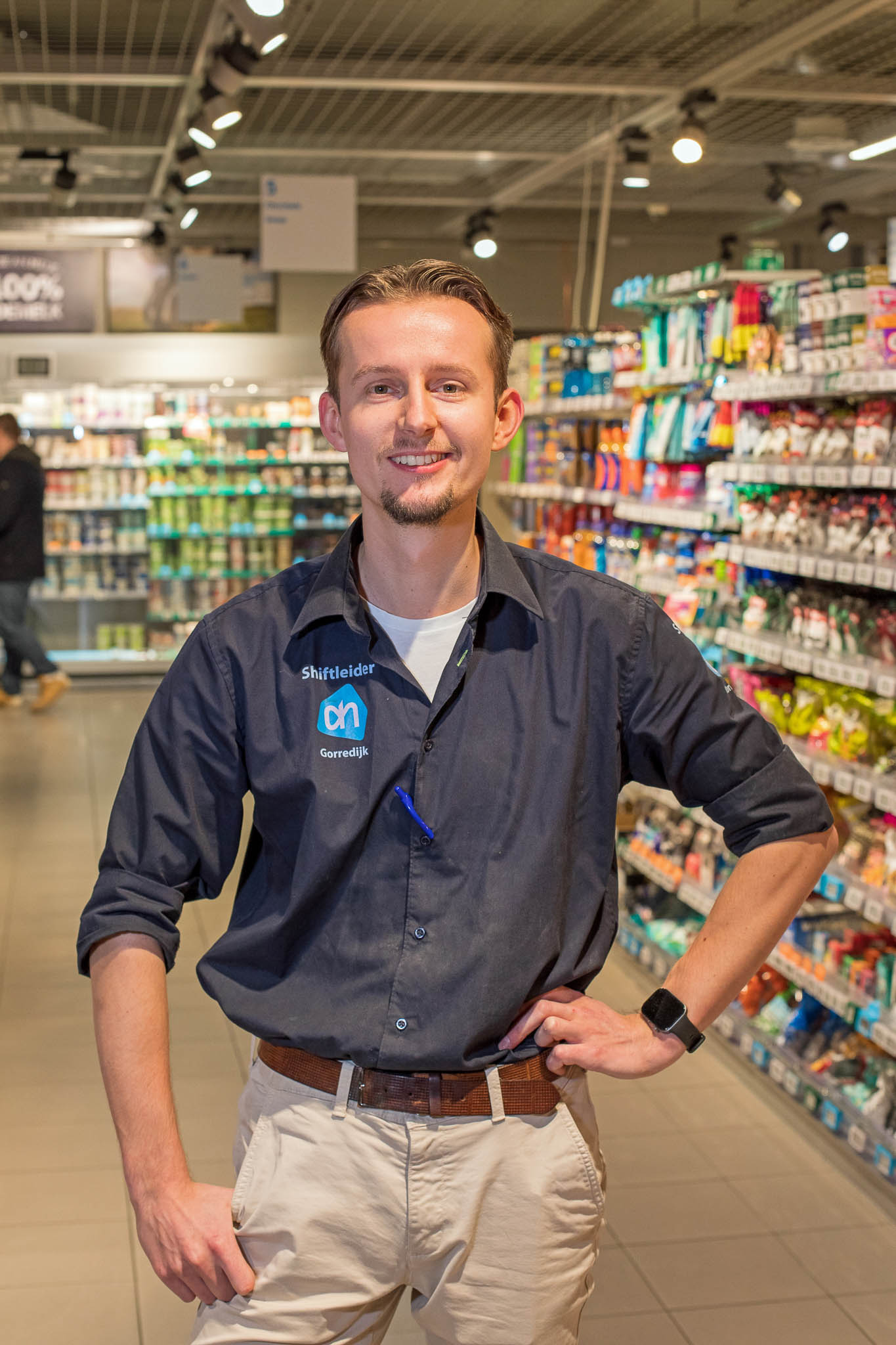 Lachende man met donker overhemd en beige broek staat in een supermarktgang met schappen vol producten.