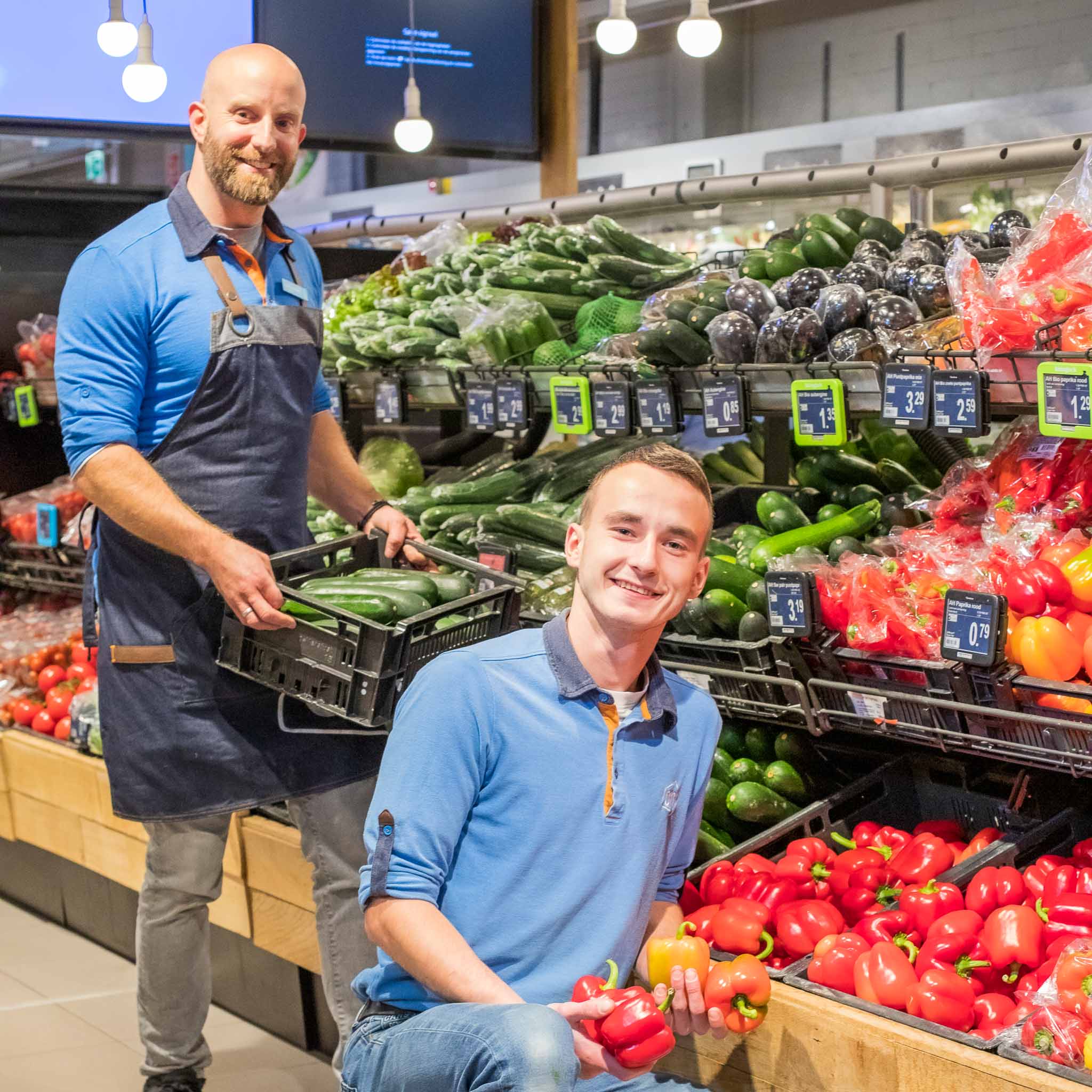 Twee supermarktmedewerkers, een staat met een krat komkommers en de ander knielt terwijl hij rode en gele paprika's vasthoudt bij een groentenafdeling.