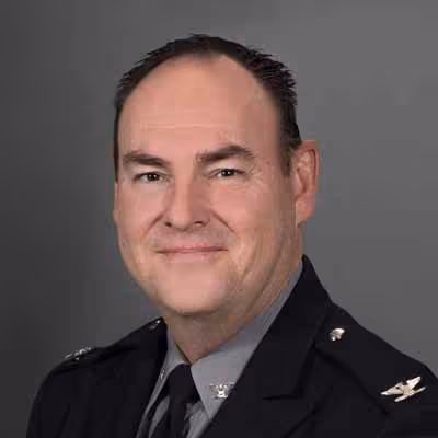 Man in a black police uniform with shoulder pins and tie, smiling against gray background.