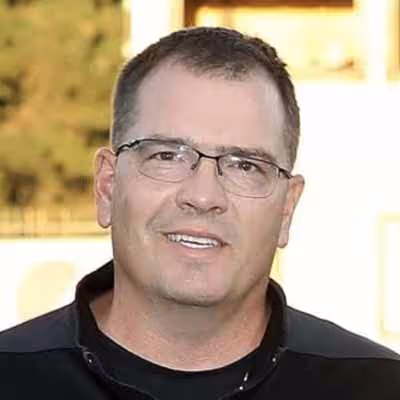 Man with short dark hair and glasses, wearing a black shirt, smiling outdoors.