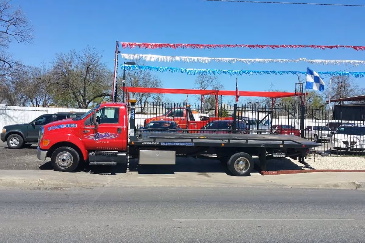 Red flatbed tow truck parked on street with colorful flags and cars in the background.