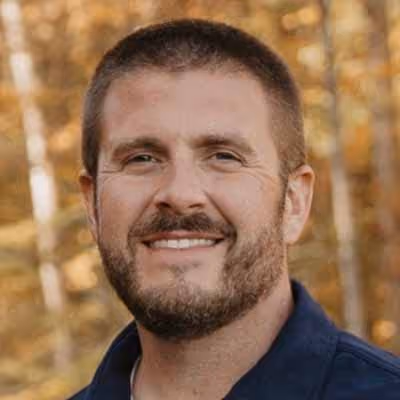 Smiling man with short hair and beard standing outdoors with blurred autumn trees in background.