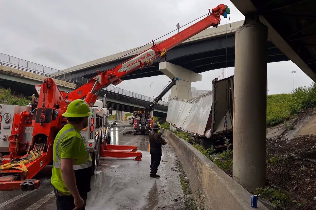 Tow trucks and workers lifting a damaged semi truck under highway overpasses on a rainy day.