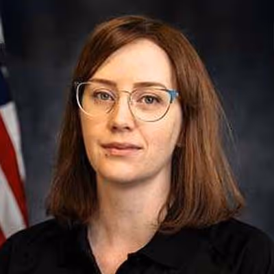 Woman with shoulder-length auburn hair and glasses wearing a black top against a dark background.