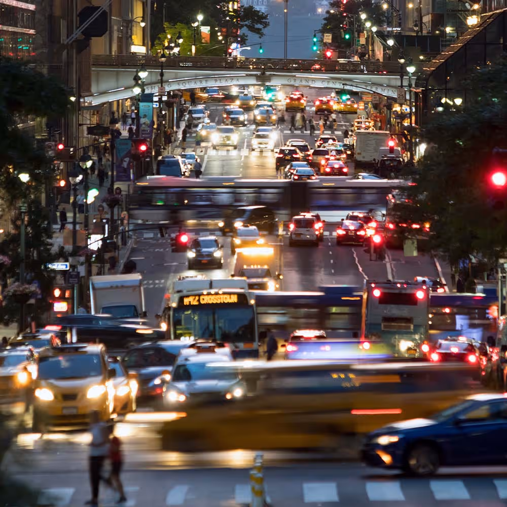 Busy city street at dusk with heavy traffic, buses, cars, and pedestrians near a bridge.