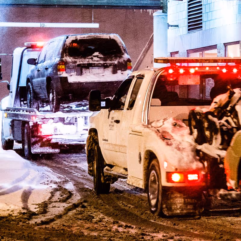 Tow truck hauling a snow-covered SUV on a snowy urban street at night with lights flashing.