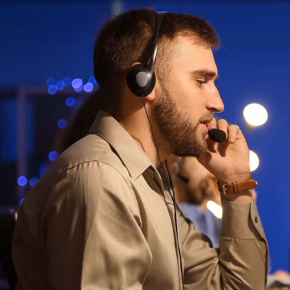 Bearded man wearing headset speaking into microphone in a dimly lit room with blurred lights.