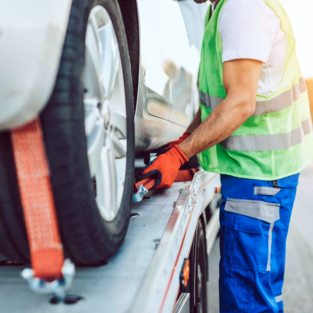 Tow truck driver in safety vest and gloves securing a car wheel with a red strap on a tow truck.