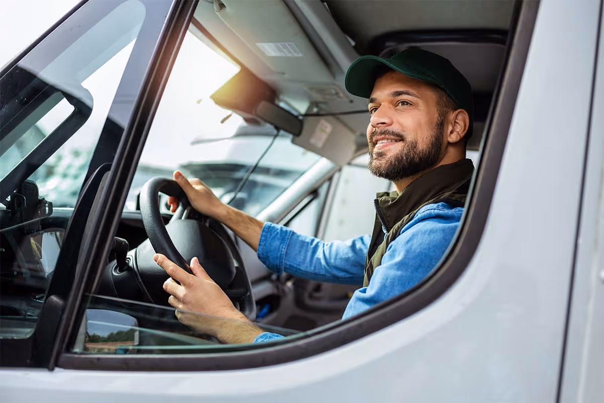 Smiling man wearing cap driving a van, visible through the open driver's side window.