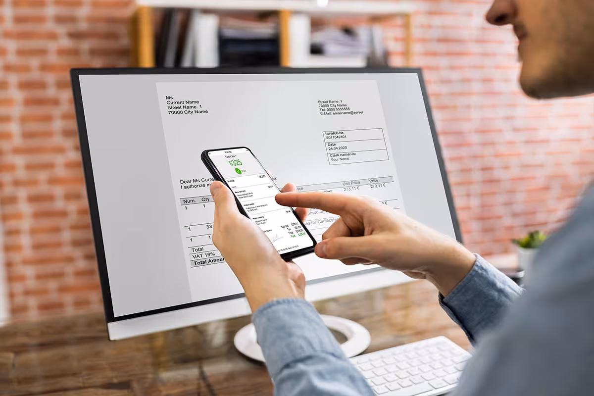 Person holding smartphone displaying a $325 payment with an invoice displayed on a computer screen.