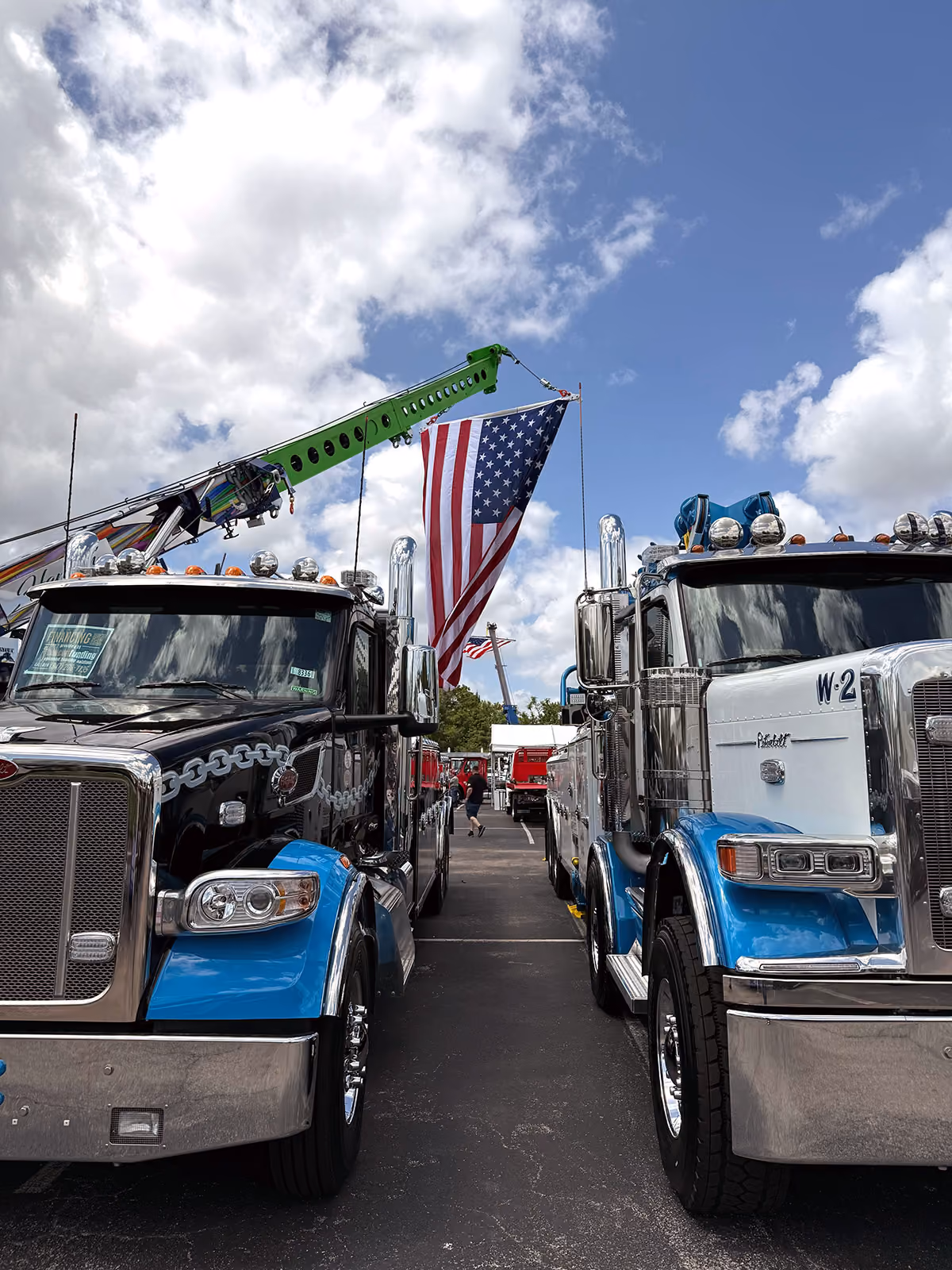 Two blue and black semi trucks with a large American flag hanging between them under a partly cloudy sky.