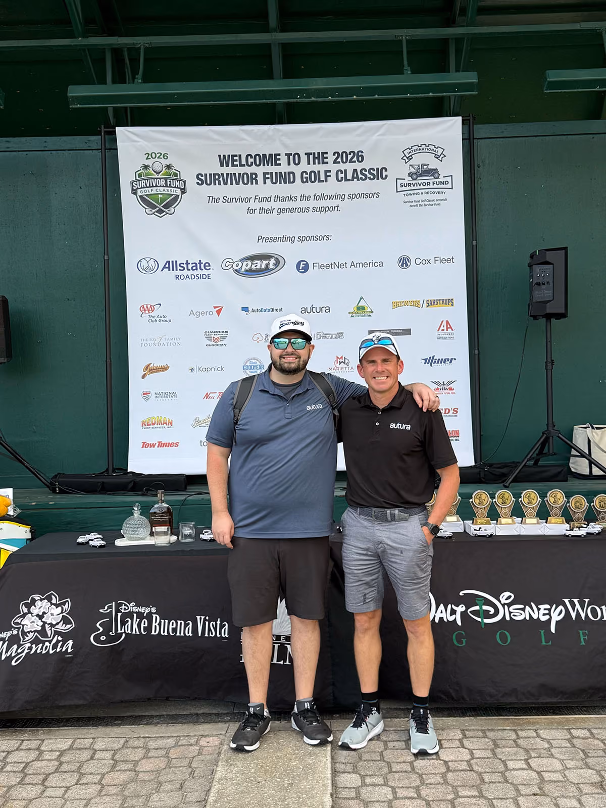 Two men in golf attire stand side by side in front of a 2026 Survivor Fund Golf Classic sponsor banner.