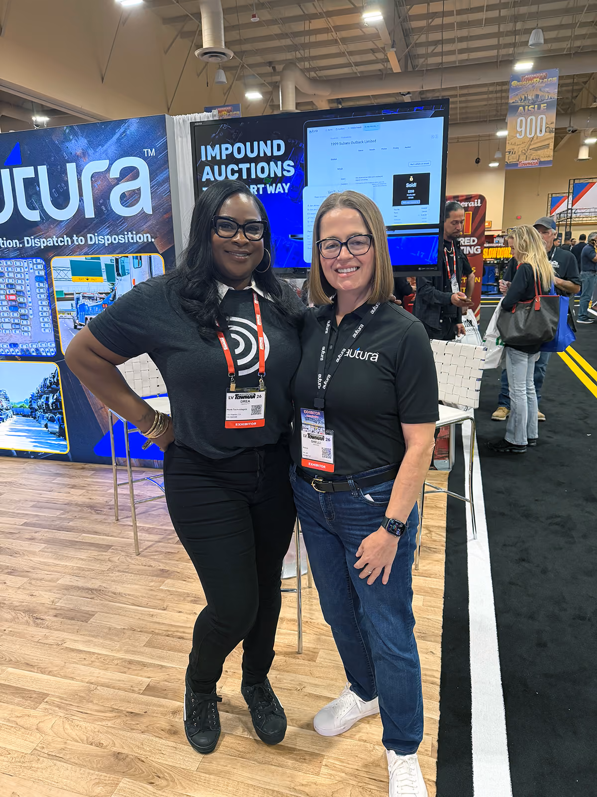 Two women wearing Autura shirts and exhibitor badges smiling at a trade show booth.