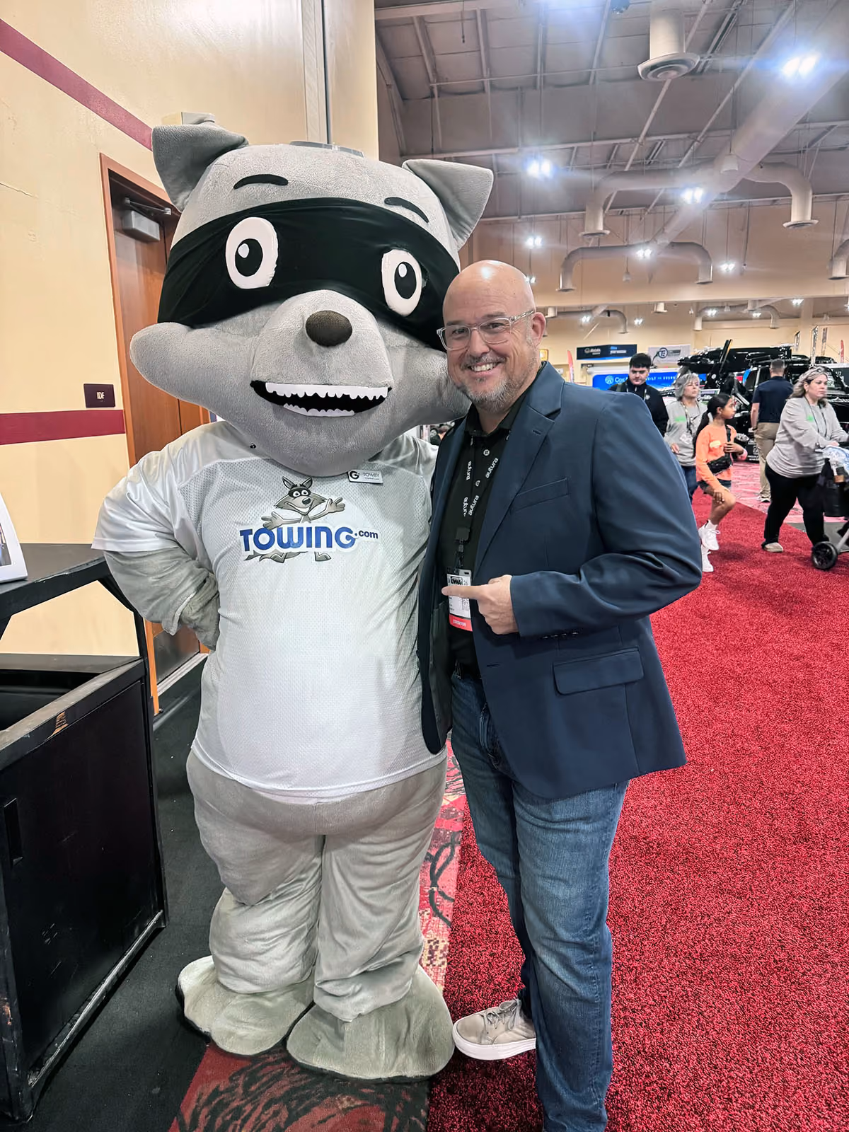 Man in glasses and blazer smiles and points at a raccoon mascot wearing a Towing.com jersey.