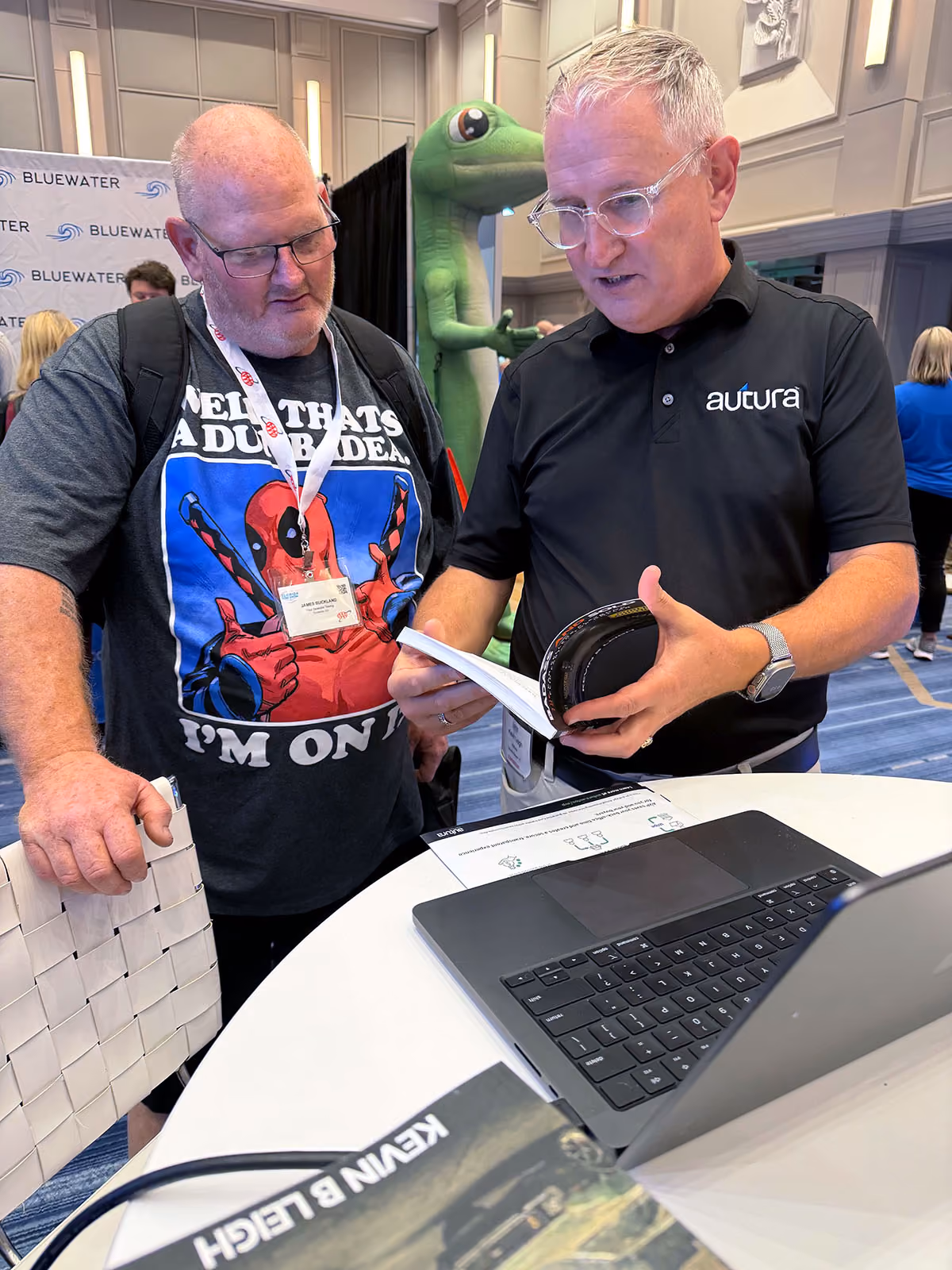 Two men at a conference table reviewing a booklet with a laptop and a green lizard mascot behind them.