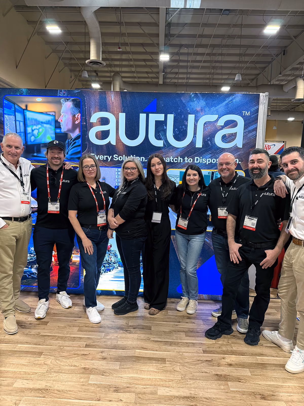 Group of nine people posing in front of an Autura booth at a trade show or event.