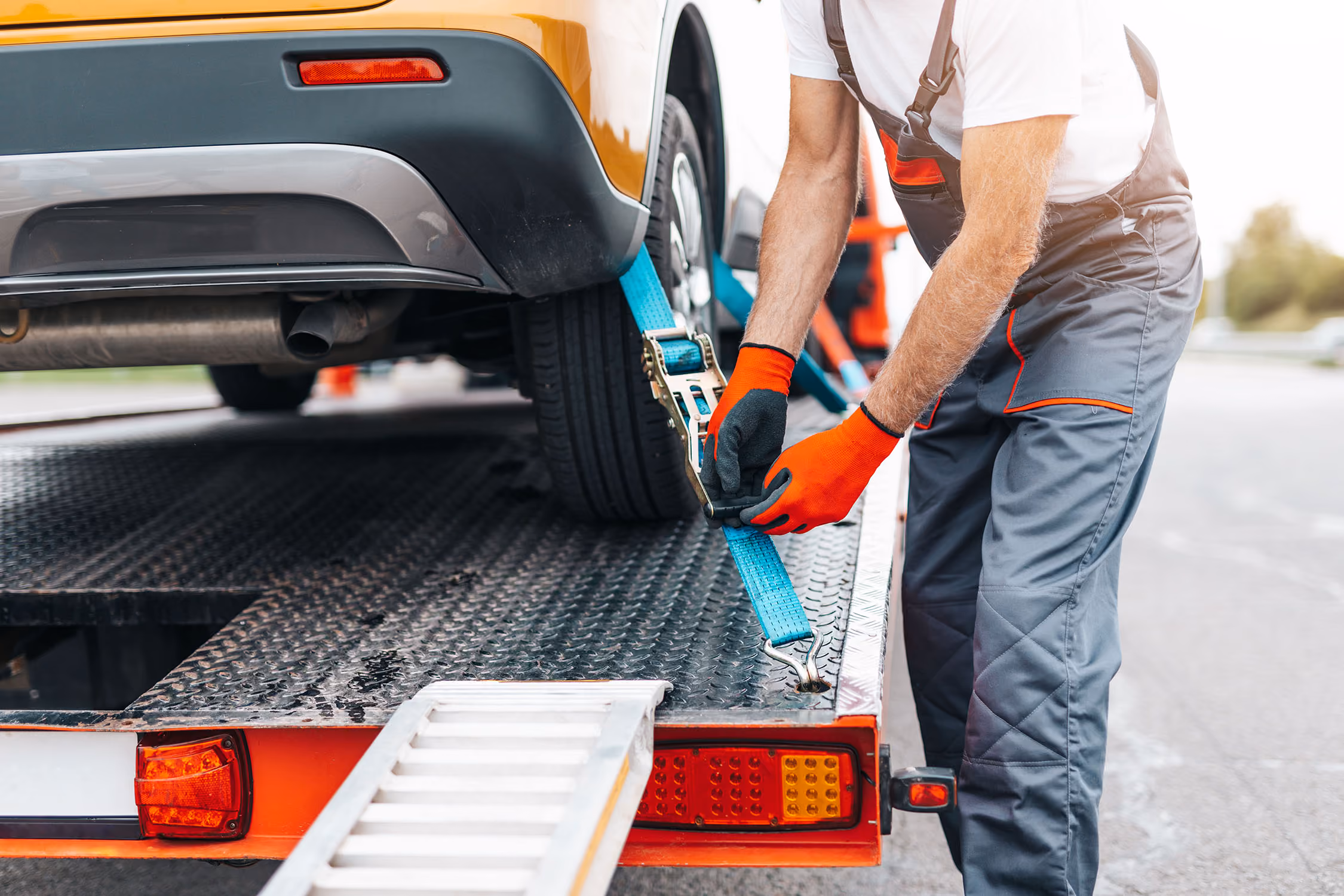Tow truck worker fastening blue strap securing yellow car on flatbed truck.