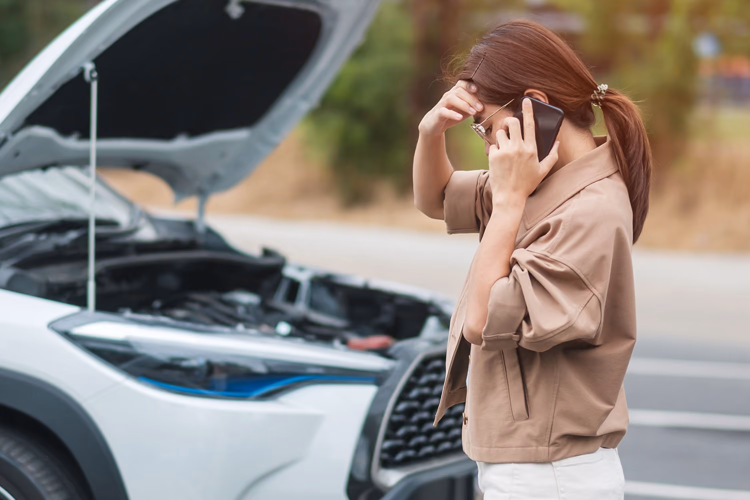 Woman distressed on the phone standing in front of a white broken down car on the side of the road