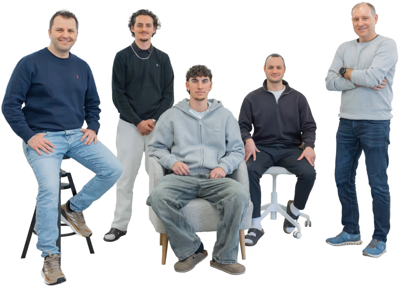 Fünf Männer in legerer Kleidung, drei sitzend und zwei stehend, vor neutralem Hintergrund.