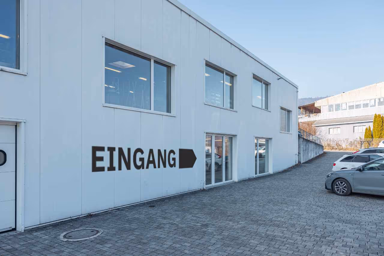 Weisses Bürogebäude mit grossen Fenstern und mehreren parkierenden Autos auf gepflastertem Vorplatz.