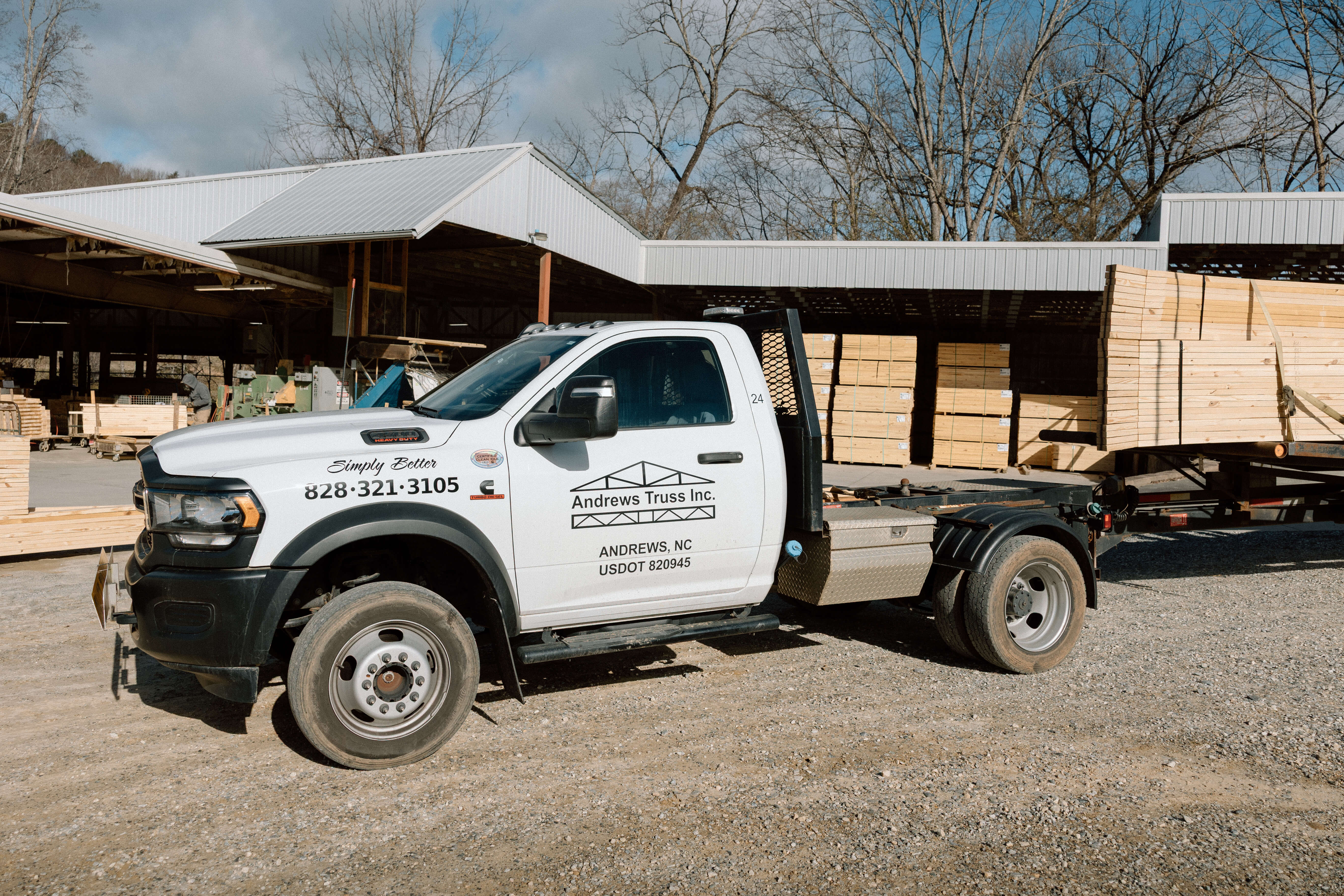 White Andrews Truss Inc. truck parked outside a lumber yard with stacks of wooden boards and a trailer loaded with lumber.