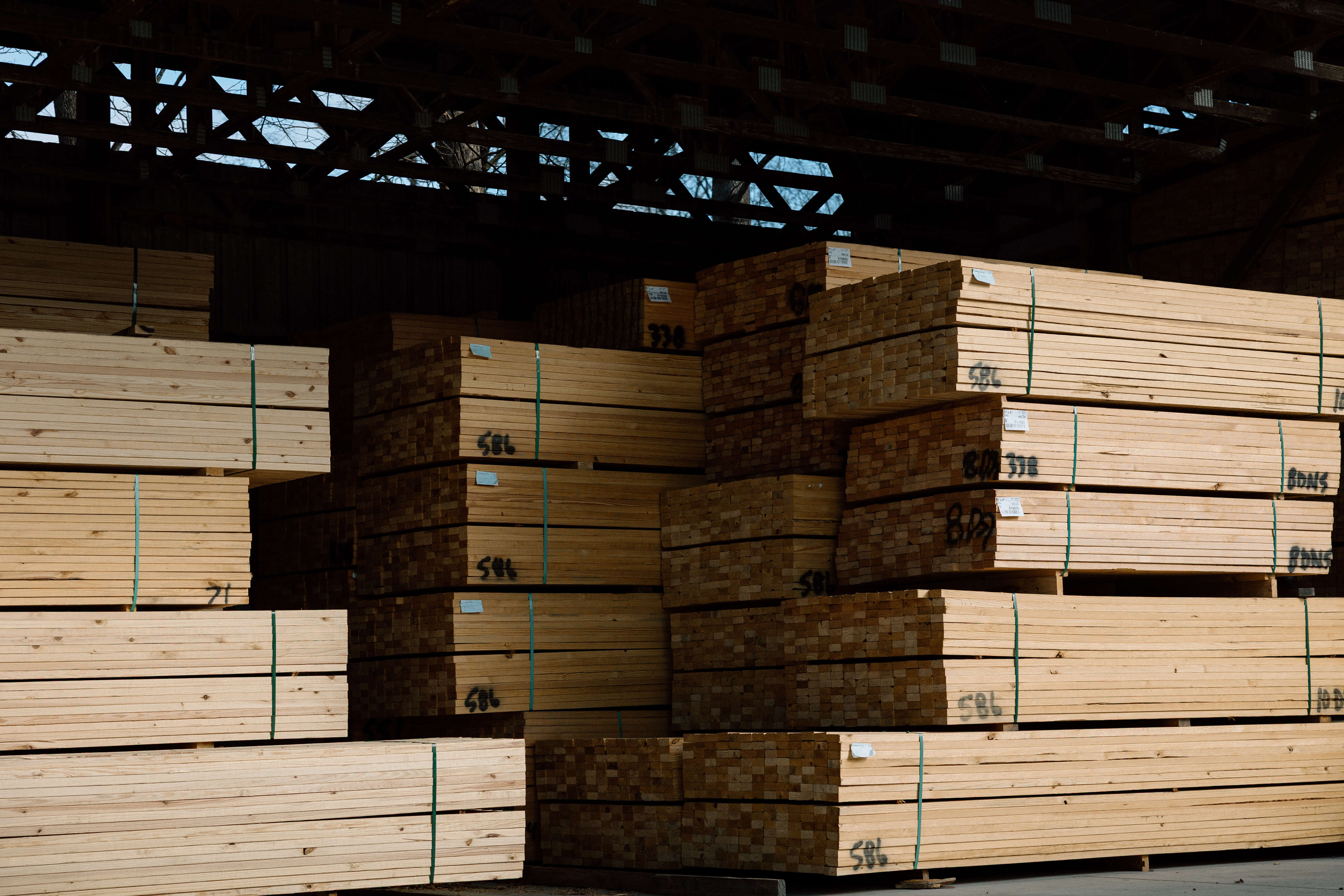 Stacks of bundled wooden planks stored inside a warehouse with exposed roof beams.