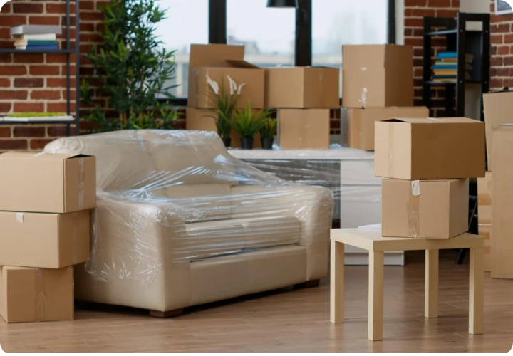 Living room with beige armchairs wrapped in plastic and multiple cardboard moving boxes on floor and table.