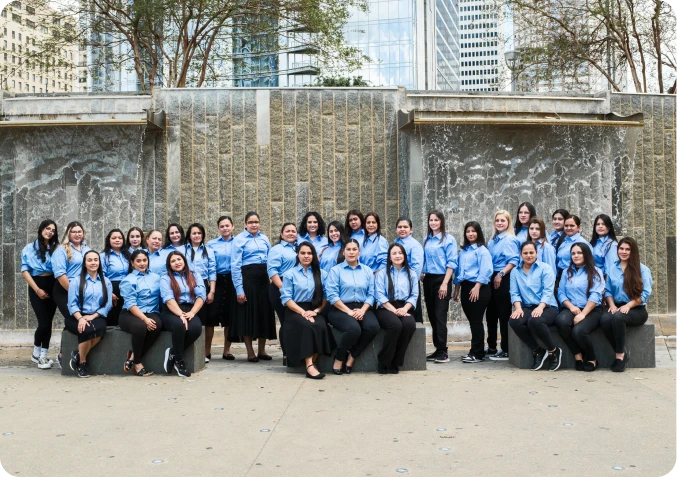 Group of 27 women wearing matching blue shirts posing outdoors in front of a stone wall with water features.