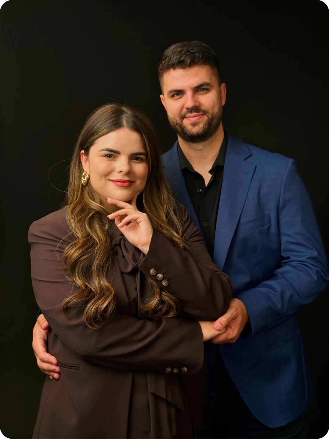 Smiling couple posing with the man hugging the woman from behind against a black background.
