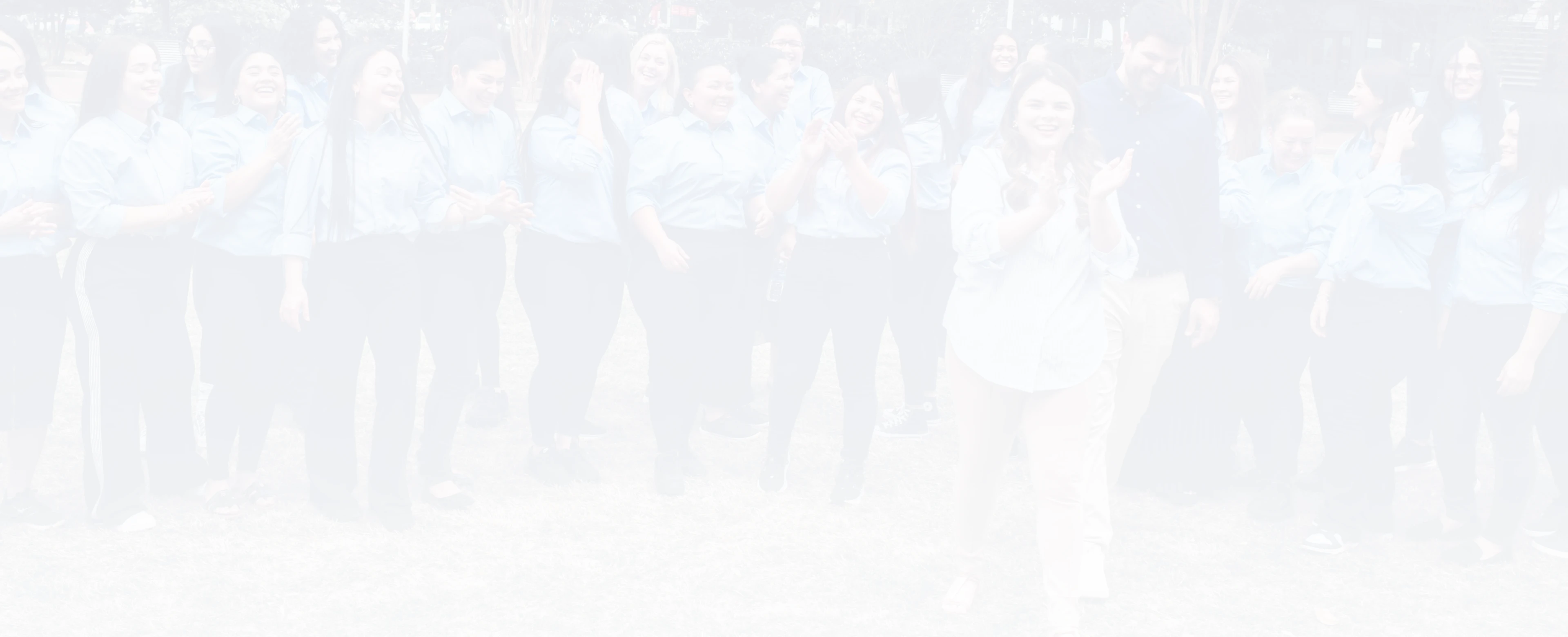 Group of smiling young women and one man standing outdoors on grass, many wearing blue shirts and clapping.