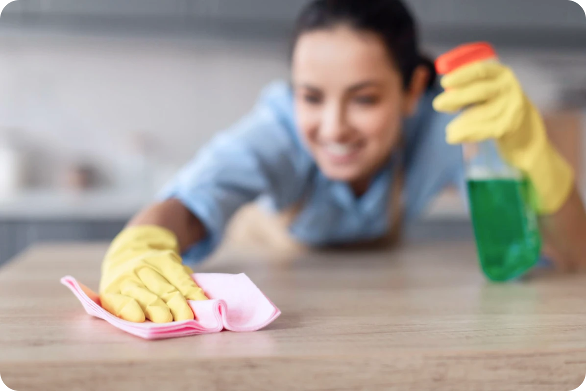 Person wearing yellow rubber gloves cleaning a wooden surface with a pink cloth and green spray bottle.
