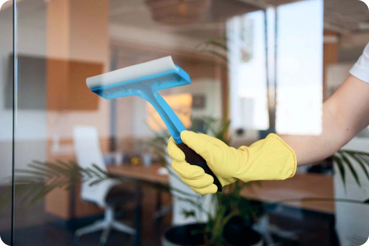 Hand wearing a yellow glove holding a blue squeegee cleaning a glass window in an office setting.