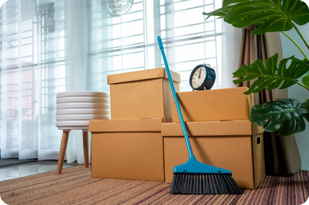 Cardboard moving boxes stacked next to a blue broom and an alarm clock on a striped carpet near a window with sheer curtains.