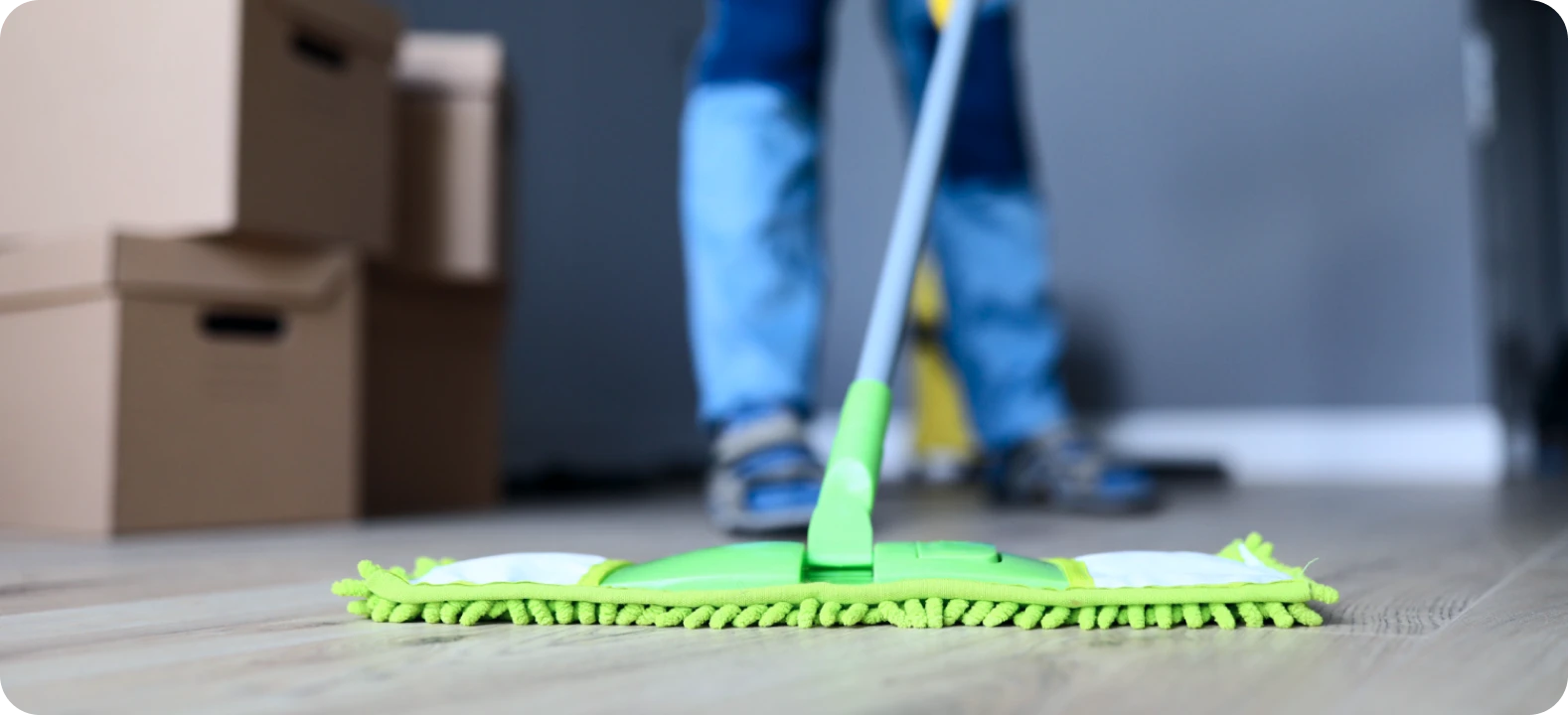 Person mopping light-colored wooden floor with a green flat mop near cardboard moving boxes.