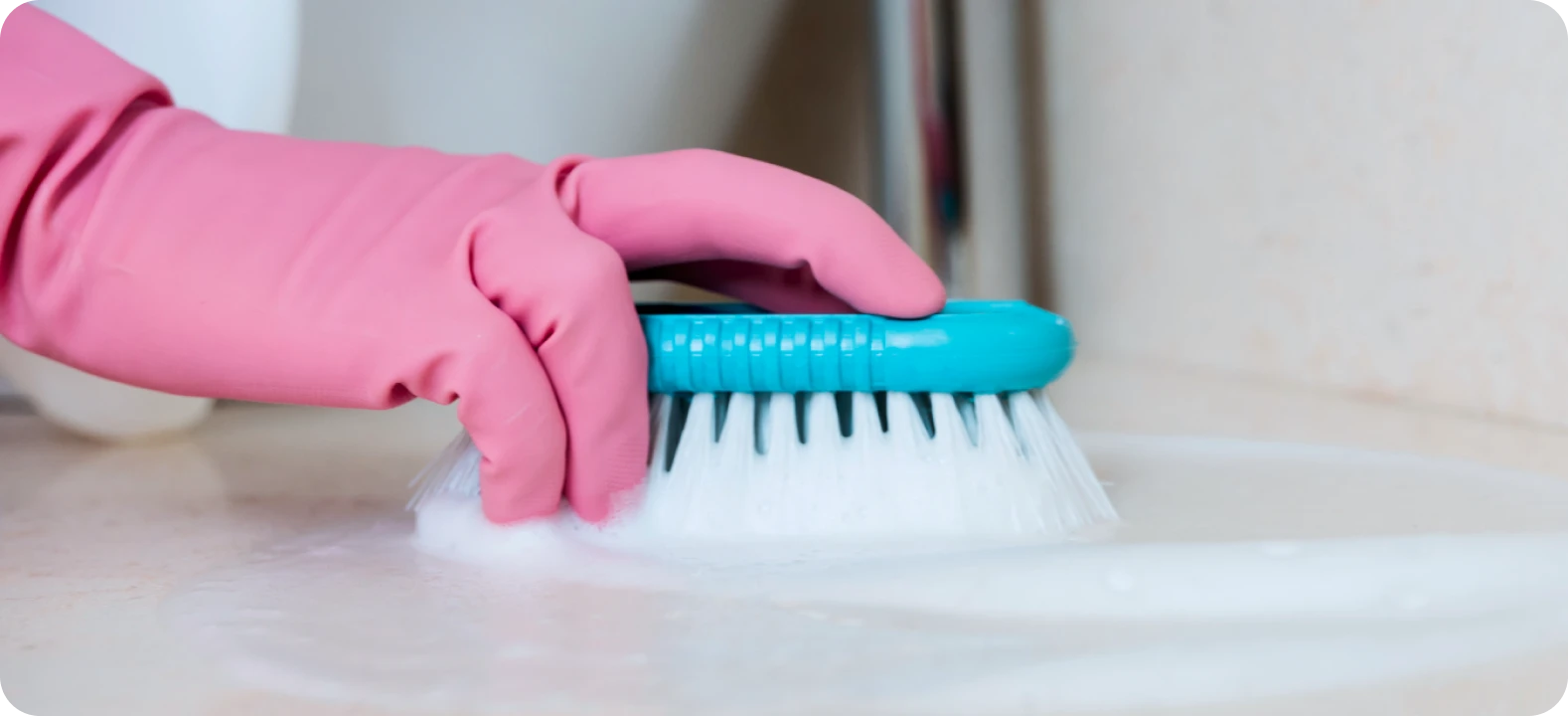 Hand in pink glove scrubbing a surface with a blue brush and white foam.