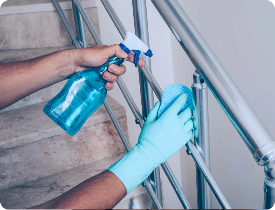 Person wearing blue gloves cleaning a metal stair railing with a sponge and spray bottle.