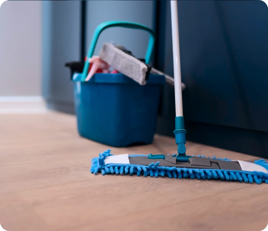 Blue mop on wooden floor with a blue bucket containing cleaning supplies in the background.