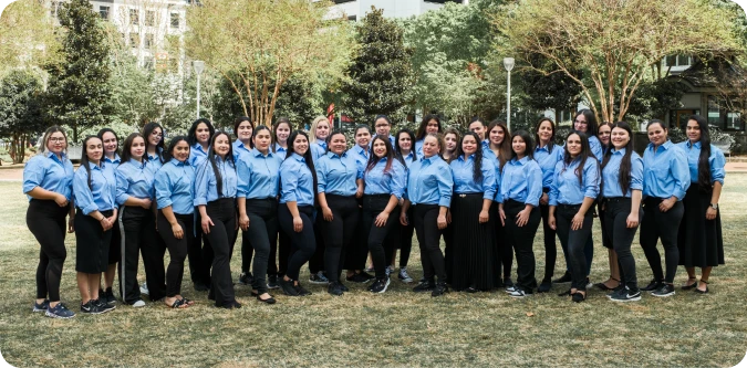 Group of 27 people standing outdoors on grass, all wearing blue shirts and dark pants or skirts, with trees and buildings in the background.