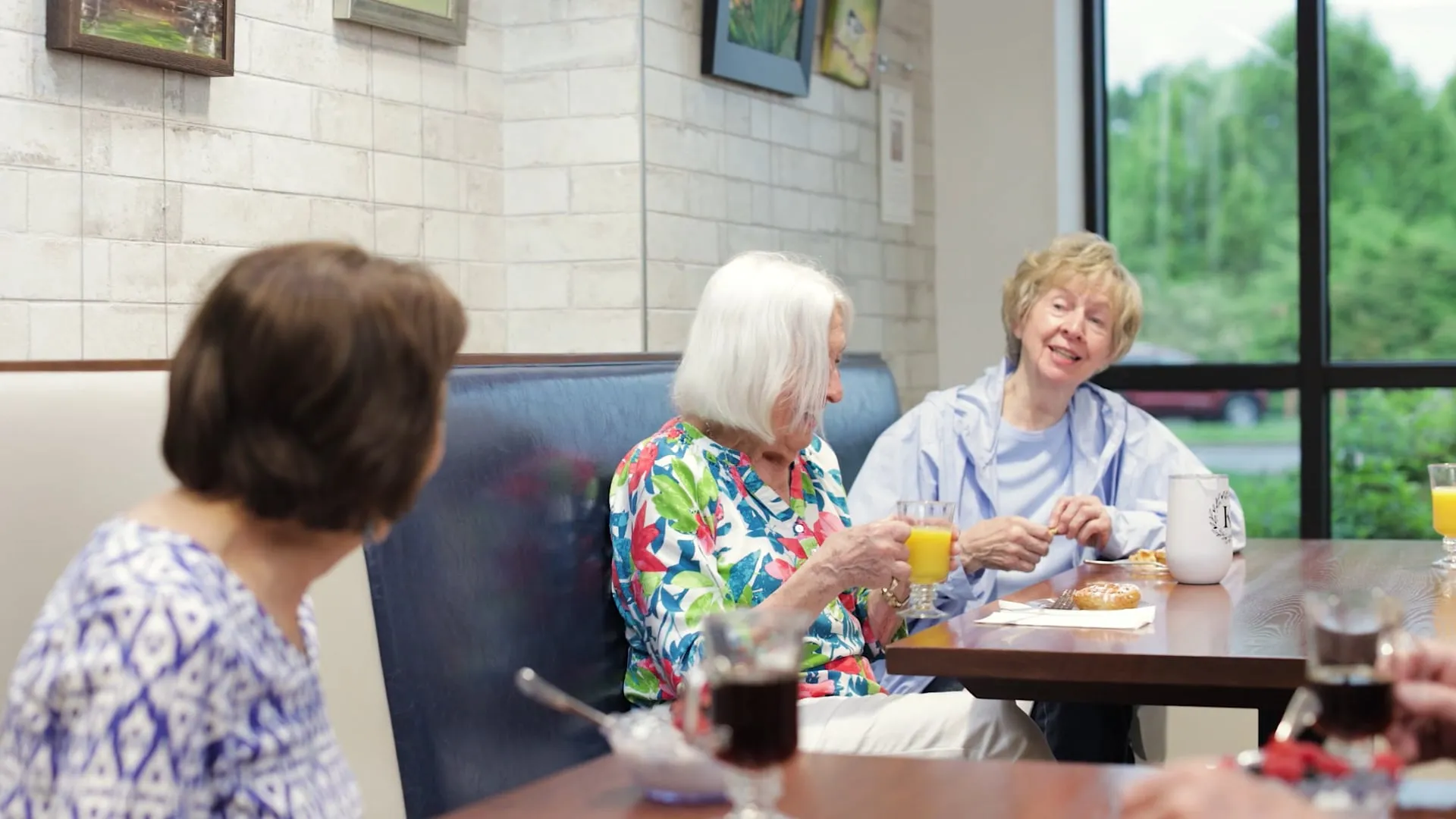 Three older women sit together at a table, talking and drinking in a casual setting.
