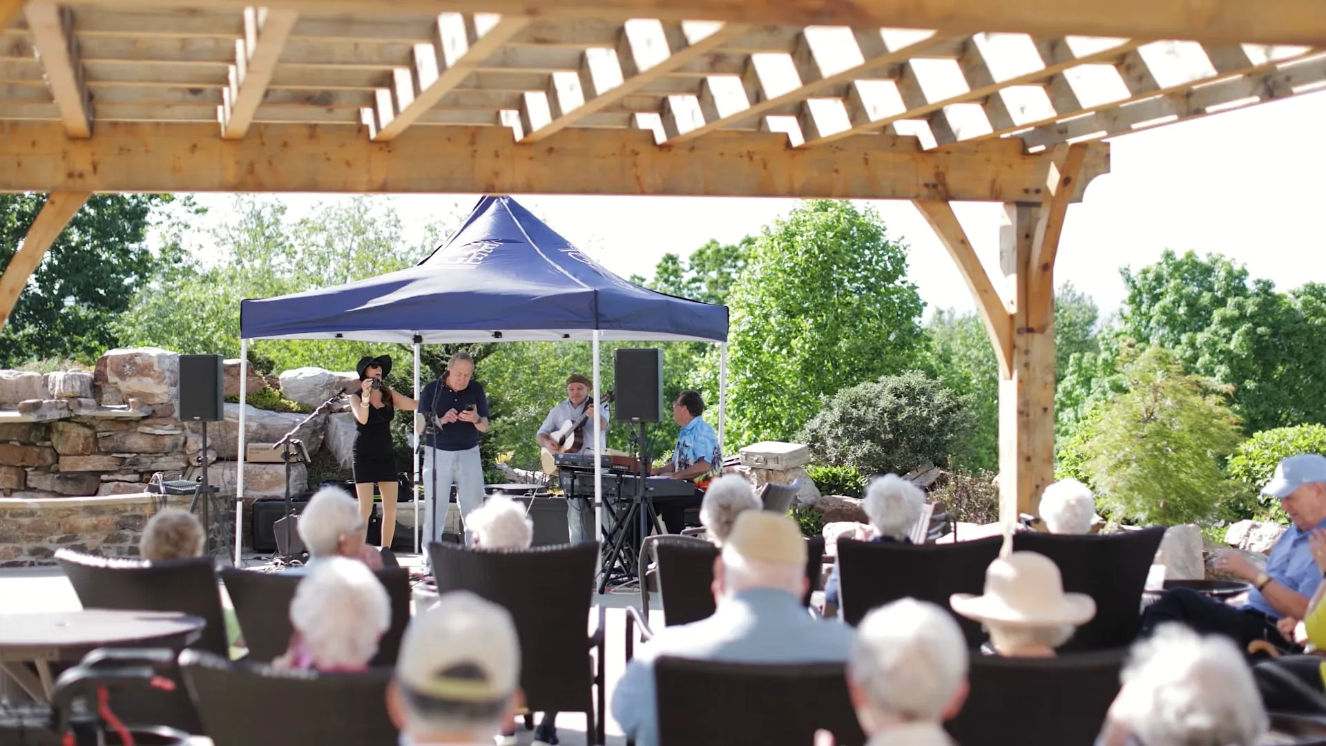 A band performs under a blue tent for an audience seated outdoors.